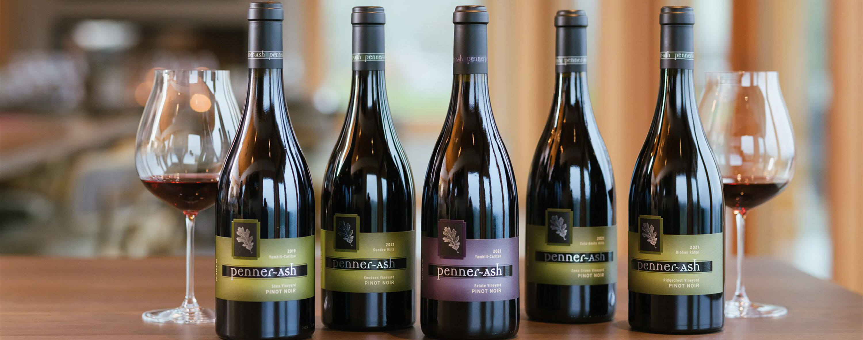 5 Penner-Ash Single Vineyard Pinot Noirs on a table between two glasses of red wine.